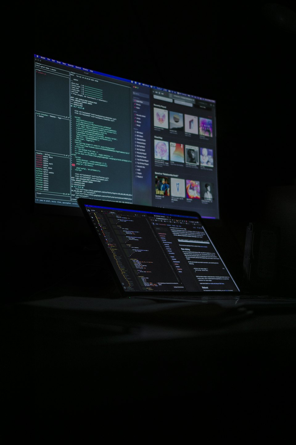 Notebook a monitor zobrazujúci zložitý kód v tmavej miestnosti.